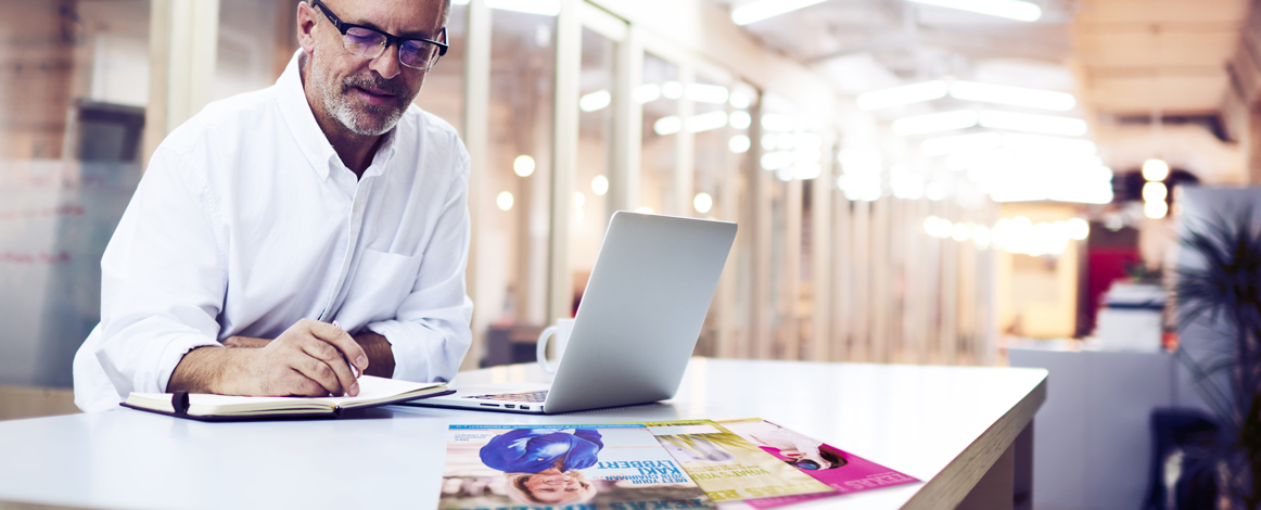 White older man with black-framed glasses sitting at a table in front of a computer, writing on a notepad with three issues of Texas REALTOR® magazine on the table
